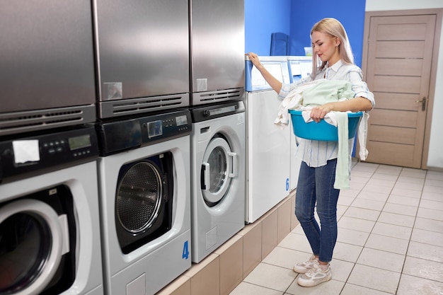 Woman standing alone with dirty clothes in the self serviced laundry with dryer machines. female in casual wear stands holding basin with clothes.