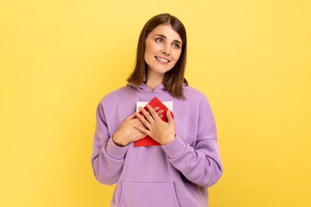 Woman standing embracing envelope and looking away with pleasant smile and enjoying