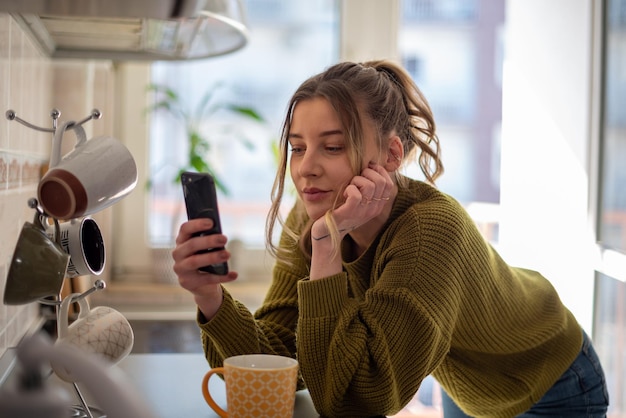 Woman standing in the kitchen and drinking cup of tea while using smartphone