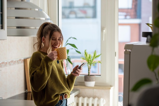 Woman standing in the kitchen and drinking cup of tea while using smartphone