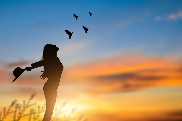 Woman standing on the meadow alone holding both arms up and\
holding a hat looking up at the sky.