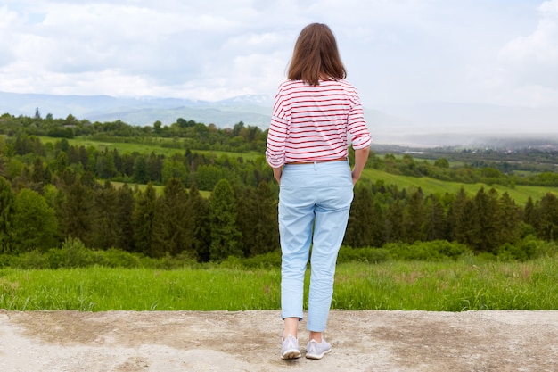 Photo woman standing in mountain, panoramic view of mountain range, trees. green grass. blue sky, female wearing casual trousers and white t shirt