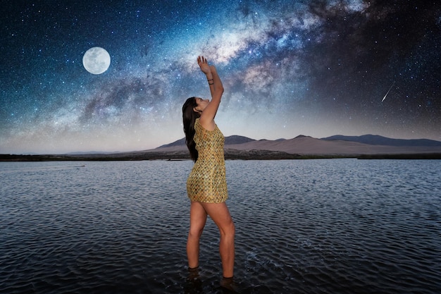 Foto donna in piedi nell'acqua che fissa il cielo stellato con la via lattea e lo sfondo della luna