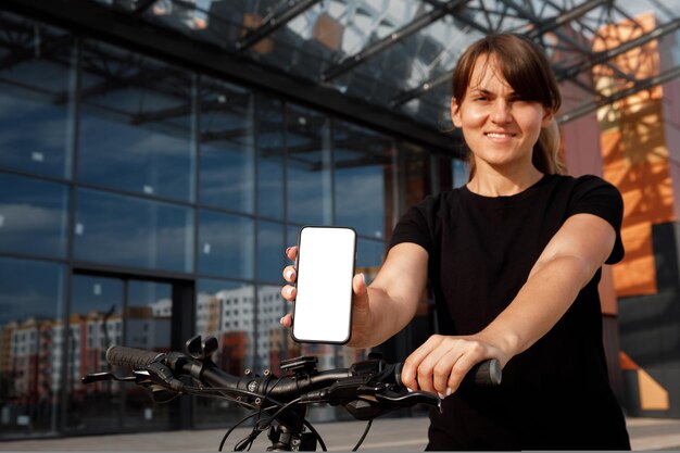 自転車で立って、空白の白い画面で携帯電話を表示している女性