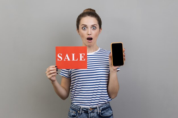 Photo woman standing with open mouth holding sale card and cell phone with empty display