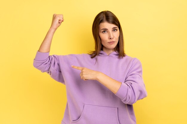 Woman standing with strong gesture and looking with serious expression pointing at her biceps