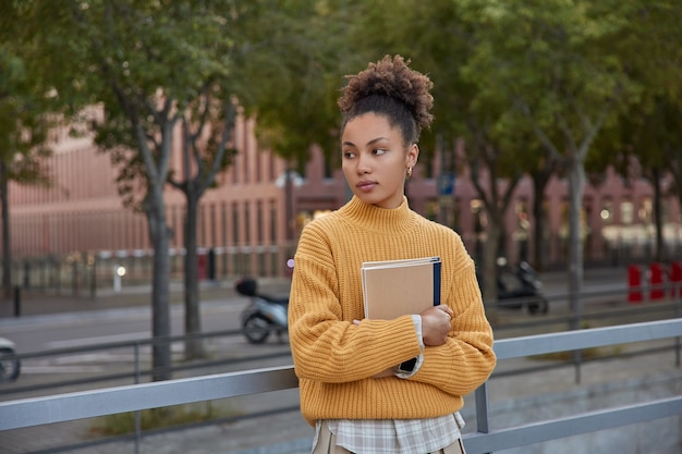 woman stands against street background holds diary or notebook for writing notes wears knitted yellow sweater focused away spends free time for learning in city during daytime