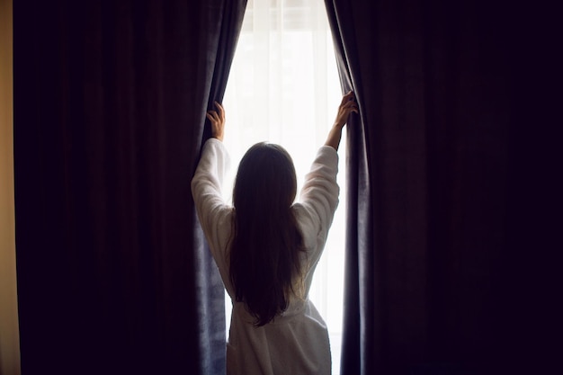 Woman stands in a bathrobe at the window and opens the curtain in the hotel