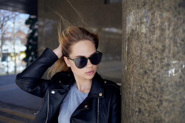 A woman stands under a bridge in the street wearing sunglasses and a leather jacket