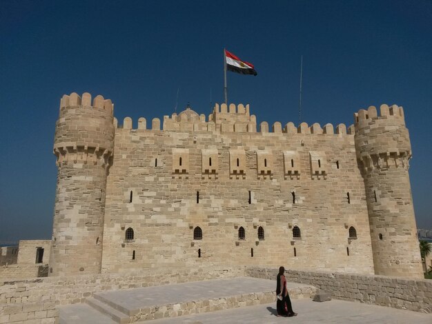 A woman stands in front of a castle with a flag on the top.