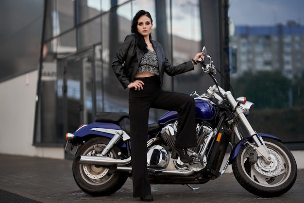 woman stands in front of a motorcycle in front of a glass building in a parking lot near a shopping center.