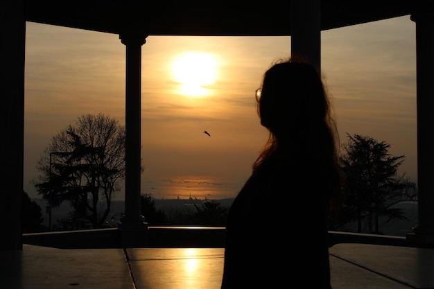 A woman stands in front of a window with the sun setting behind her.