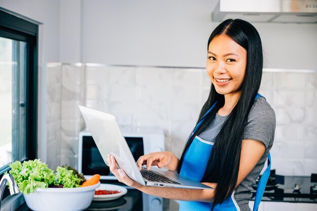 A woman stands in the kitchen happily cooking while using a laptop for video tutorials