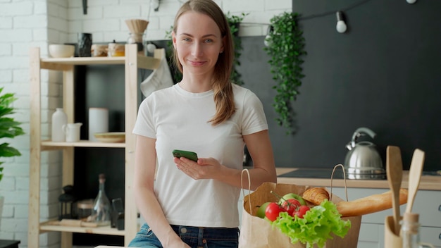 Woman stands in the kitchen near a paper bag full of fresh food and uses a smartphone app to deliver it to the supermarket