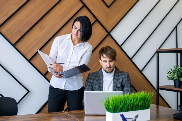 A woman stands next to a man working on a laptop making notes in a folder