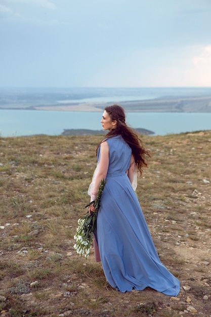 woman stands on a mountain cliff in a blue long dress in summer