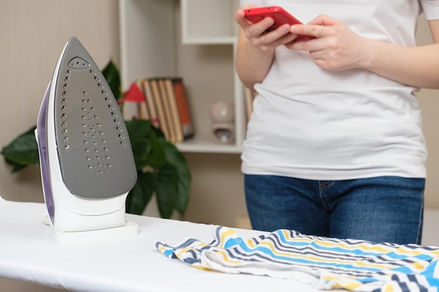 A woman stands near an ironing board with an iron and looks at the screen of her phone Tips for caring for clothes