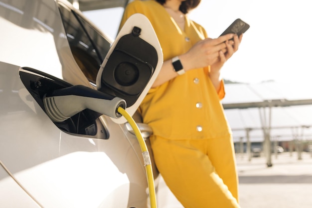 Woman stands near recharging electric car with mobile phone vehicle charging at public station