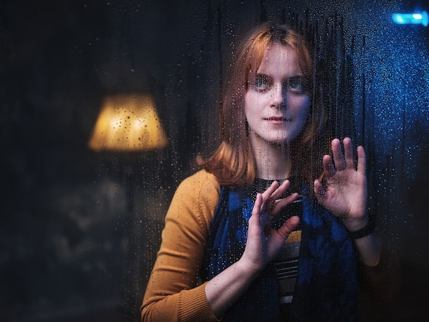 A woman stands behind a rainy window