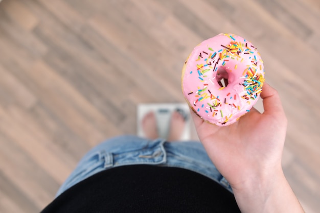 Una donna si trova sulla bilancia, tiene in mano una ciambella, il concetto di dieta, aumento di peso, perdita di peso, alimentazione corretta, dolce.