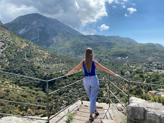 A woman stands on a stone staircase overlooking a mountain valley.