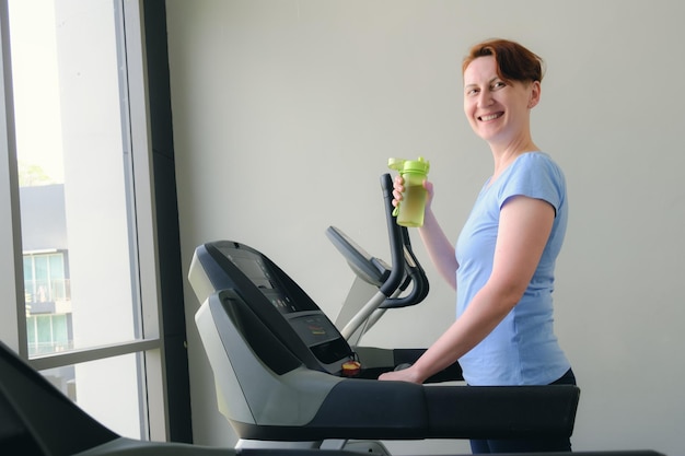 Woman stands on a treadmill in the gym hold sports bottle with water and smiles