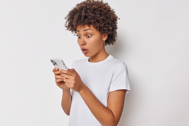 woman stares at smartphone astonished to read impressive article wears casual t shirt isolated on white gazes frightened in device screen.