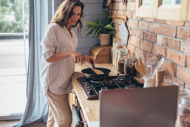 Woman stirring food cooking in a frying pan on the stove and looking at her laptop she reading cooking recipe or watching show while frying food