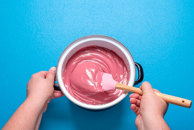 Woman stirring ruby chocolate glaze in a pot on blue background top view melted pink chocolate