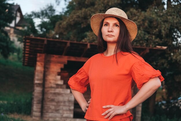 Woman in a straw hat dressed in red clothes stands on the background of a wooden house