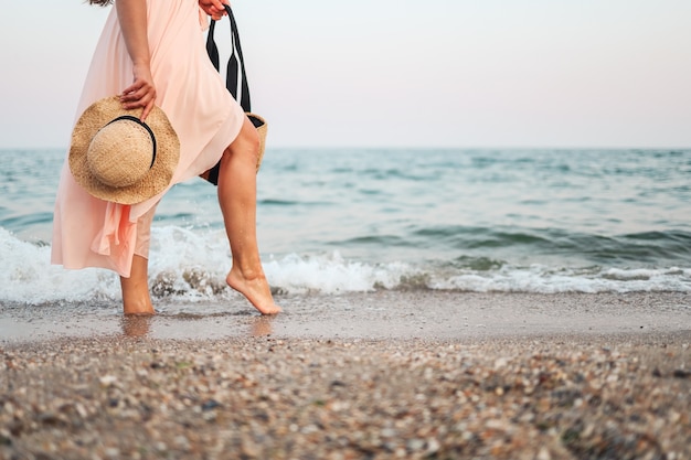 Photo woman in straw hat and pink dress on a tropical beach with brown bag.