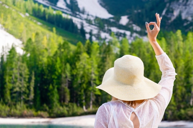 Woman in the straw hat and white dress shows the sign Ok on the background of mountains and forest