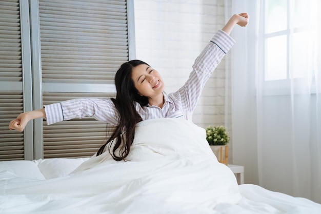 Woman stretching in bedroom after wake up. young asian korean beautiful lady sitting in comfort white bed smiling enjoy morning with sunlight. happy charming female raised arms and hands joyful face.