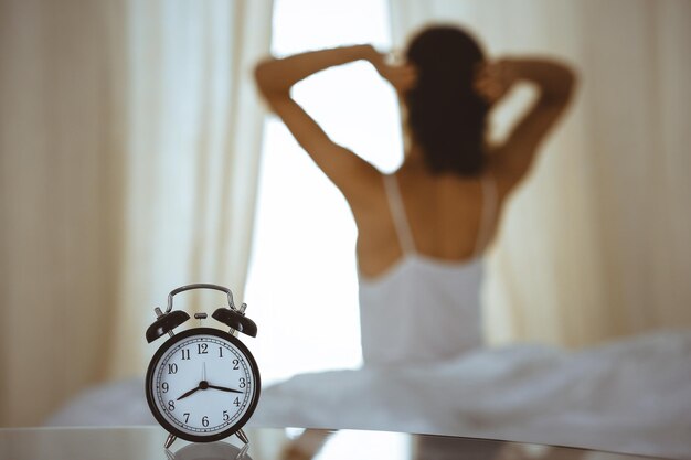 Woman stretching hands in bed after wake up, sun flare . Brunette entering a day happy and relaxed after good night sleep and back view. Concept of a new day and joyful weekend.