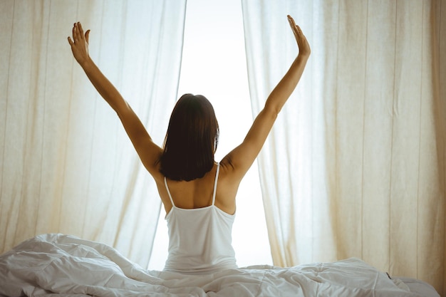 Woman stretching hands in bed after wake up, sun flare . Brunette entering a day happy and relaxed after good night sleep and back view. Concept of a new day and joyful weekend.