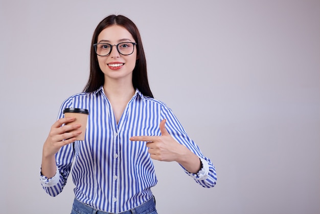 woman in a striped shirt and black pc glasses stands with a brown cup of coffee in her hand on gray. The lady calmly looks straight. Smile.