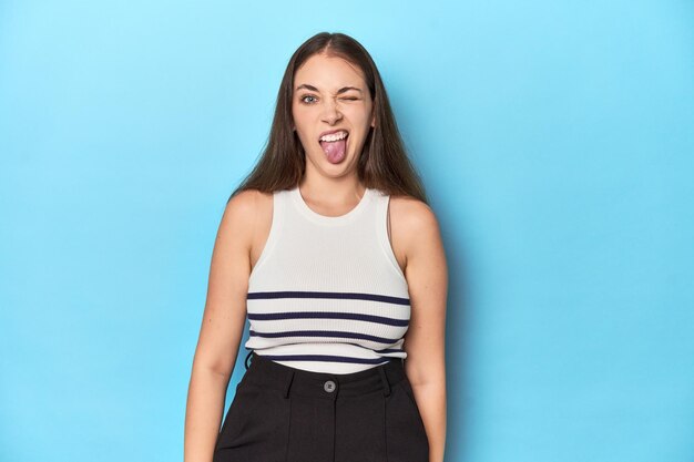 Woman in a striped top posing on a blue studio backdrop funny and friendly sticking out tongue