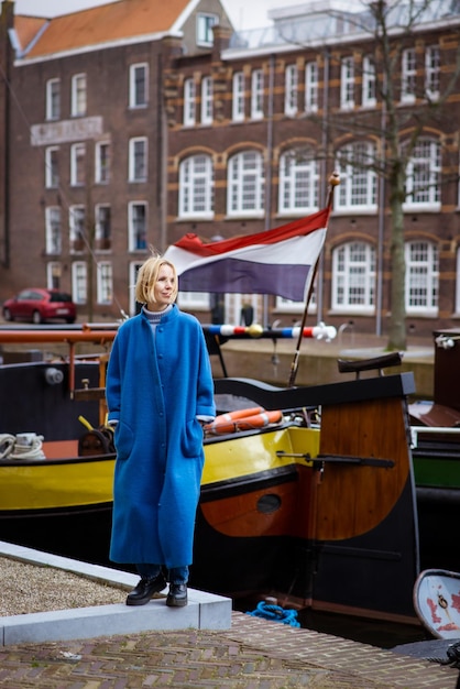 Foto una donna con un elegante cappotto blu su un canale con navi e una bandiera francese che sventola sentimenti di passione per i viaggi e l'avventura zona residenziale nei paesi bassi