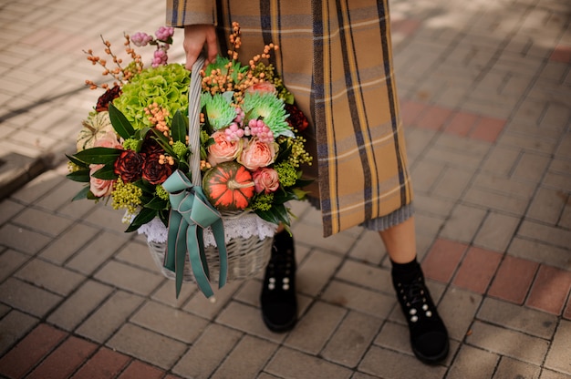 美しい秋の花の組成の大きな枝編み細工品バスケットを保持しているスタイリッシュな格子縞のコートを着た女性