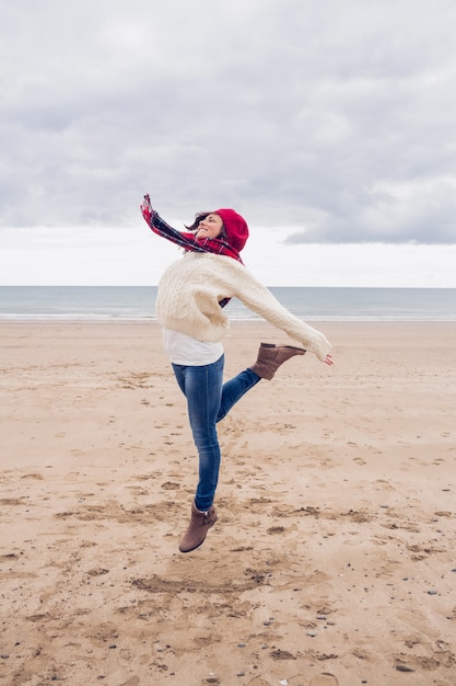 Женщина в стильной теплой одежде прыгает на пляже