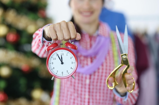 Woman stylist holds red alarm clock and scissors on background of christmas tree sewing evening