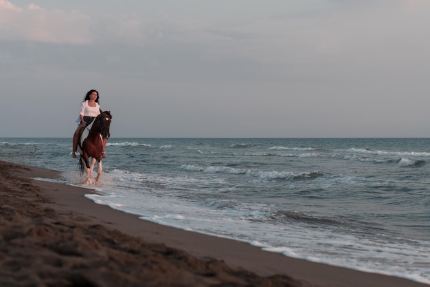 夏服を着た女性は、夕日のセレクティブ フォーカスで美しい砂浜で馬に乗って楽しんでいます