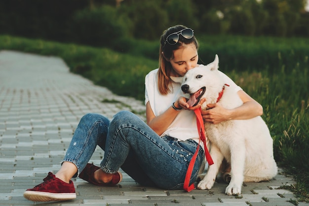 Woman in summer clothes sitting on pavement with crossed legs hugging and kissing happy white dog with open jaws looking at camera