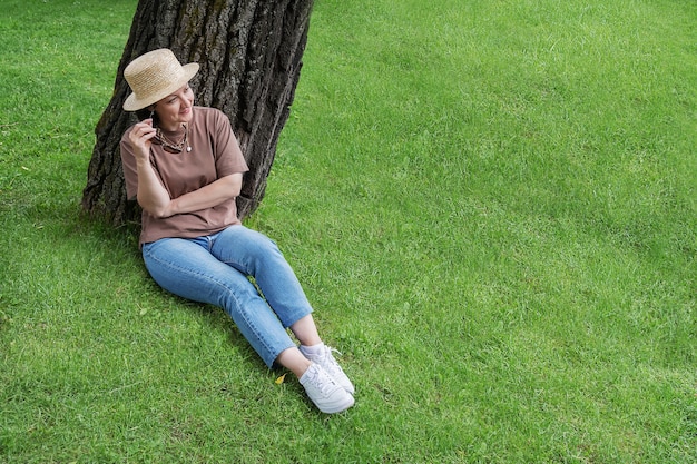 Una donna in abiti estivi e un cappello di paglia si siede vicino a un albero sul prato e sorride.