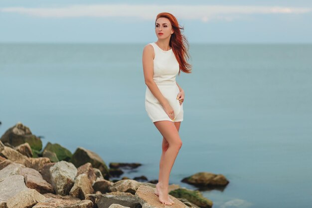 A woman in a summer dress is standing by the sea and looking away