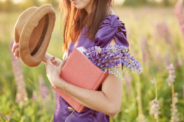 Woman in summer field with bouquet of wildflowers lupines hat
and pink notebook closeness to nature selfdiscovery concept relax
and welllbeing concept sunny day good mood concept