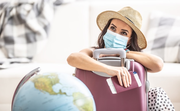 Woman in summer hat and face mask leans against the suitcase holding flight ticket and a passport thinking of traveling during pandemic.