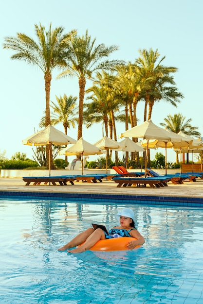 A woman in a sun hat in the pool in a swimming circle with a laptop works remotely