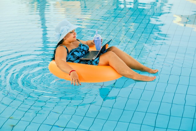 Woman in a sun hat and a swimsuit in the pool in a swimming circle works with a laptop.