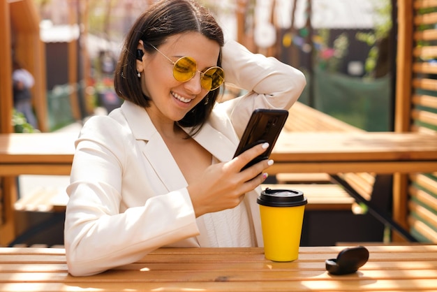 Женщина в солнцезащитных очках с маленькими наушниками в ушах смеется, глядя на смартфон
